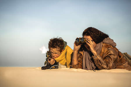 Post apocalyptic woman and boy with binoculars and gun outdoors. Desert and dead wasteland on the background. Aggressive girl warrior in shabby clothes and young boy with weapon laying a sand in ambush shooting someone. People in nuclear post-apocalypse time. Life after doomsday concept.の写真素材