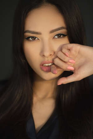 Asian woman beauty face closeup portrait. Beautiful young girl in black dress posing indoors looking at the camera. Close-up fashion portrait of Asian female model with perfect makeup isolated on a dark studio background.の写真素材