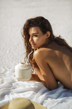 Young mixed race Asian Caucasian woman with no bikini holding coconut cocktail, laying on the beach. Gorgeous lady with nude perfect body. Women in paradise place creative concept photo.の写真素材