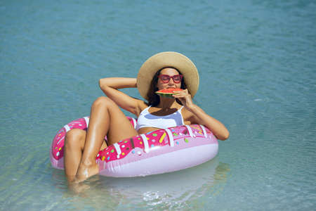 Travel and vacation concept. Young beautiful woman in bikini hat and sunglasses eating watermelon, relaxing in the sea on a rubber ring. Happy girl floating on an inflatable colorful donut with a fruit during summer sunny day. Front view of a slim lady, clear water. Copy space background.の写真素材