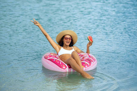 Travel and vacation concept. Young beautiful woman in bikini hat and sunglasses eating watermelon, relaxing in the sea on a rubber ring. Happy girl floating on an inflatable colorful donut with a fruit during summer sunny day. Front view of a slim lady, clear water. Copy space background.の写真素材