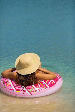 Travel and vacation concept. Young beautiful woman swimming in bikini hat and sunglasses relaxing in the sea on a rubber ring. Happy girl floating on an inflatable colorful donut with in the water in summer sunny day. Back view of a slim lady, clear water. Copy space background.の写真素材