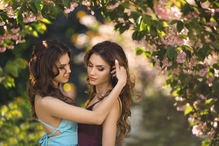 Portrait of fashionable twins models with perfect make-up and hairstyle standing near sakura blossoming tree. Spring beauty concept. Portrait shot of two gorgeous women in evening dresses with the perfect look, posing resting on nature.の写真素材