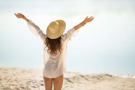 Romantic woman rising hands feeling free, relaxing on a beach against a beautiful sea bokeh background. Tanned girl in fashionable trendy boho style rear view. Travel vacation, summer outdoor pleasure concept.の写真素材