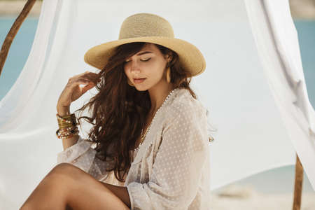 Portrait of attractive brunette woman sitting on a deserted beach under the handmade tent. Gorgeous slim mixed race Asian Caucasian woman a white dress, hat, jewelry spending holidays on the sea shore. Tanned beautiful model in bohemian chic clothing resting with closed eyes on the shore in sunny day.の写真素材