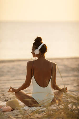 Woman meditation on the nature. Young beautiful woman in bikini meditating and listening to calm music in yoga lotus pose on deserted tropical beach outdoors. Tanned Mixed race Caucasian Asian slim girl sitting with headphones dressed in swimsuit relaxing listening sounds for relax and meditation at sunset, enjoying her vacations. Rear viewの写真素材