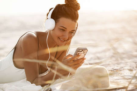 Young beautiful woman portrait in white bikini using a smartphone and listening to the music on the tropical beach outdoors. Tanned Mixed race Caucasian Asian slim girl laying with headphones and phone dressed in swimsuit relaxing at sunset, enjoying her vacations at sea. Copy space sea and sky backgroundの写真素材