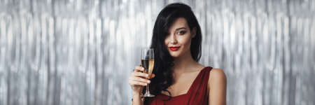 Stunning celebrating woman in the red dress. Beautiful brunette girl portrait with perfect makeup and hairstyle against holiday glowing bokeh background holding a glass wine. Fashionable luxury and festive young woman. Banner panorama with copy space for textの写真素材
