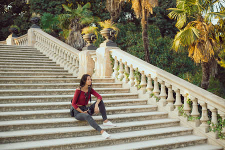 Young girl with backpack outdoors. Happy woman dressed in casual clothes sitting in a city park. Holidays and tourism concept.の写真素材
