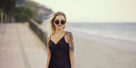 Blonde Woman in Black Dress Outdoors Against Seashore on the Background During Summer Beach Holidaysの写真素材
