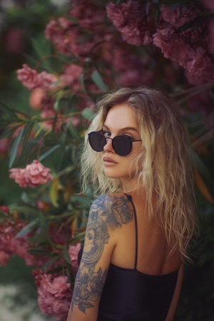 Tattooed Blonde Woman in Sunglasses Posing Near Pink Flowersの写真素材