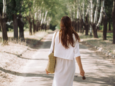 Back View of Woman Strolling on a Sunlit Tree-Lined Pathの写真素材