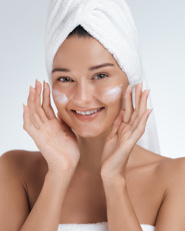 Happy Woman with Towel on Head Applying Skincare Cream with a Radiant Smileの写真素材