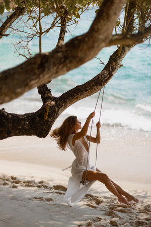 Elegant Woman Swinging on Tree by Beach in Sheer Dressの写真素材