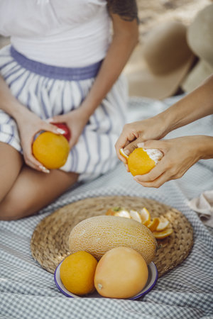 Hands Peeling Orange During Outdoor Picnic With Melons and Summer Fruits
