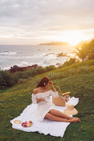 Elegant Woman Enjoying Sunset Picnic By The Ocean With Tea And Flowersの写真素材