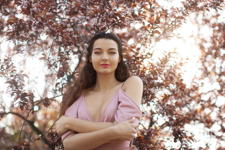 Confident Woman in Pink Dress Standing Amid Spring Blossoms at Sunsetの写真素材