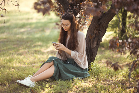 Stylish Woman Texting on Smartphone While Relaxing in Springtime Parkの写真素材