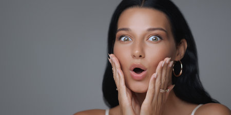 Woman Expressing Surprise with Wide Eyes and Hands on Face in Studioの写真素材