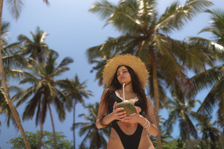 Woman In Swimsuit And Straw Hat Holding Coconut Under Tropical Palm Treesの写真素材