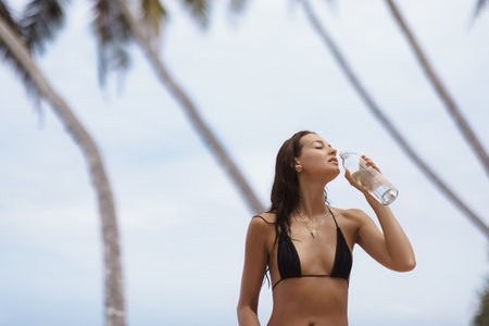 Tanned Asian Model Drinking Water From Glass Bottle At Beach On Sunny Dayの写真素材