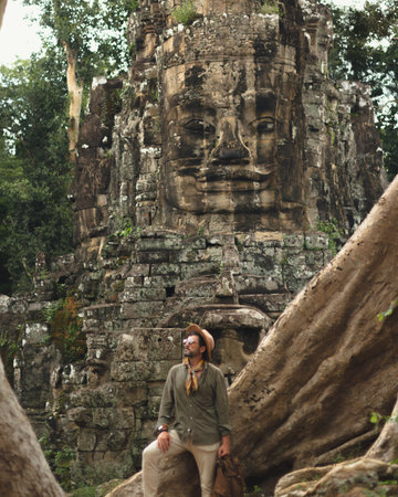 Male Explorer Standing Before Ancient Bayon Temple Face in Cambodia Jungleの写真素材