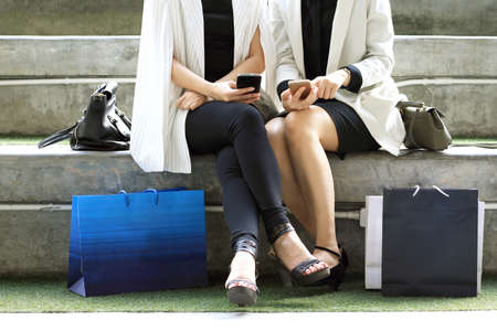 Women sitting outdoors with shopping bags and using smart phones, Concepts about lifestyle,shopping,technology and friendship.の写真素材