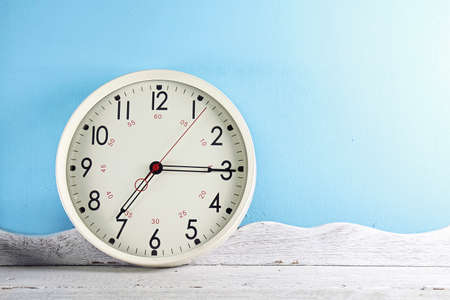 Vintage white wall clock on wooden table in front of blue background.
