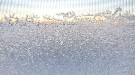 Frozen frost crystals forming delicate natural patterns on window glass at sunrise. Seasonal winter...
