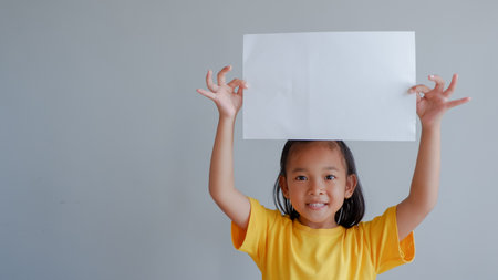 Portrait of a young asian little girl holding white paper blank sign over gray backgroundの写真素材