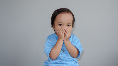 cute asian toddler wearing blue tshirt closing his mouth by hands over greyの写真素材