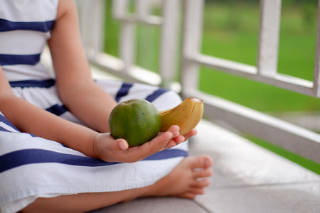 Little girl sitting on the balcony and holding banana and tangerineの写真素材
