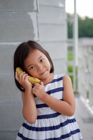 Portrait of cute little asian girl doing phone cal with banana on the house balconyの写真素材