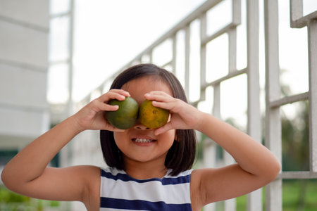 cute little asian child holding two orange fruit in front of her eyesの写真素材