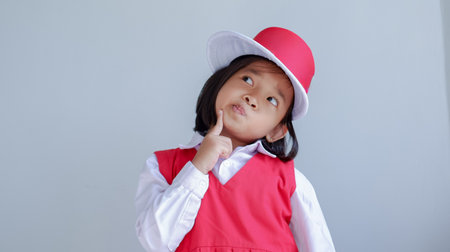 kindergarten student wearing red school uniform and hat, Indonesian kindergarten. Hand on chin looks pensively, thinking pose.の写真素材
