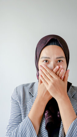 indoor portrait of beautiful Indonesian Muslim woman wearing hijab and formal clothes, covering mouth gesture amazed with shocked situation.の写真素材
