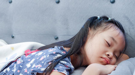 Little Asian girl with long black hair sleeping comfortably on the couch at home. Beautiful kid model lying on the tummy.の写真素材