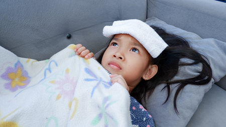 Asian child girl fever on couch with white towel on forehead. Kids healthcare conceptの写真素材