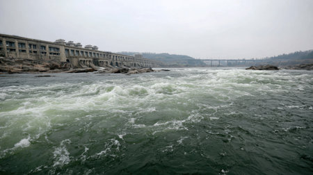 This captivating image showcases a flowing river with dynamic water movement, bordered by infrastructure in a muted, hazy atmosphere.の素材