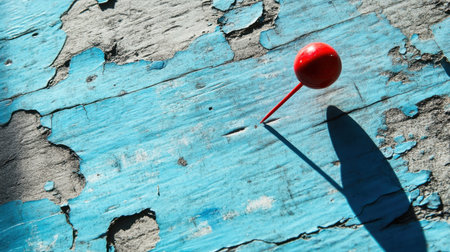 This close-up image features a bright red push pin resting on a distressed blue wooden surface. The vivid color and the texture of the wood create a striking visual, highlighted by the shadows cast by the pin. Perfect for creative projects or office settings, this image captures everyday office supplies in an artistic way.の素材