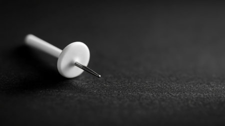 This image features a close-up view of a white push pin resting on a dark, textured surface. The moody lighting creates an elegant contrast, highlighting the pin's sharp tip and minimalist design. Perfect for themes related to organization and creativity.の素材
