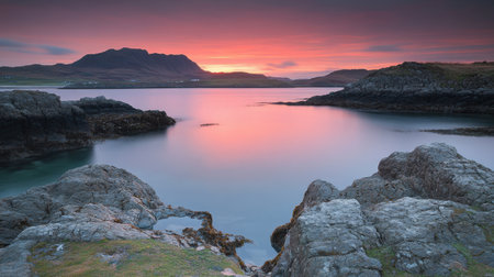 A breathtaking sunset scene captures the tranquil water reflecting vibrant hues, complemented by rugged rocks and a distant mountain range.の素材