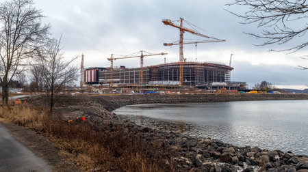 A construction site featuring multiple cranes working on a large building near a waterfront. The scene includes cloudy skies, rocky terrain, and industrial activity.の素材