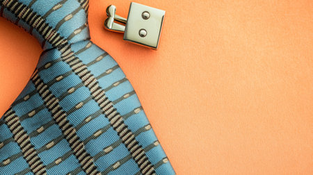 This image captures an elegant blue tie with a unique pattern, accompanied by shiny silver cufflinks, set against a vibrant orange background, perfect for fashion enthusiasts.の素材