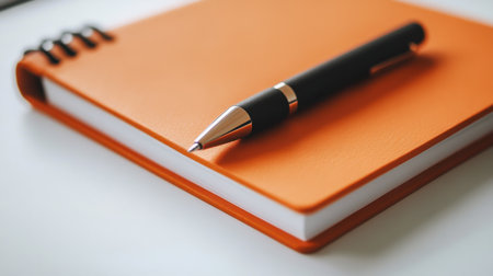 A vibrant orange notebook paired with a sleek pen, resting on a clean white surface. Ideal for writing, planning, or creative endeavors in any setting.の素材