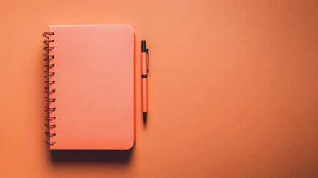 This image features a bright orange notebook accompanied by a matching pen, set against a solid orange background. Ideal for creative projects.の素材