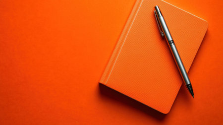 A vibrant orange notebook lies open with a sleek silver pen resting on it. This composition creates an ideal workspace for inspiration and creativity.の素材