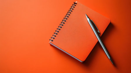 A bright orange notebook lies open with a sleek silver pen on a matching orange surface. Perfect for inspiring creativity, organization, and planning.の素材