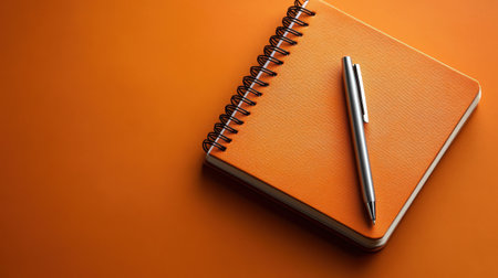 A vibrant orange notebook lies flat with a sleek silver pen resting on it, set against a matching orange background. Ideal for creative projects and academic notes.の素材