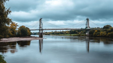 A peaceful river scene featuring a stunning suspension bridge surrounded by lush green trees, under a moody cloudy sky, exemplifying natural beauty and tranquility.の素材